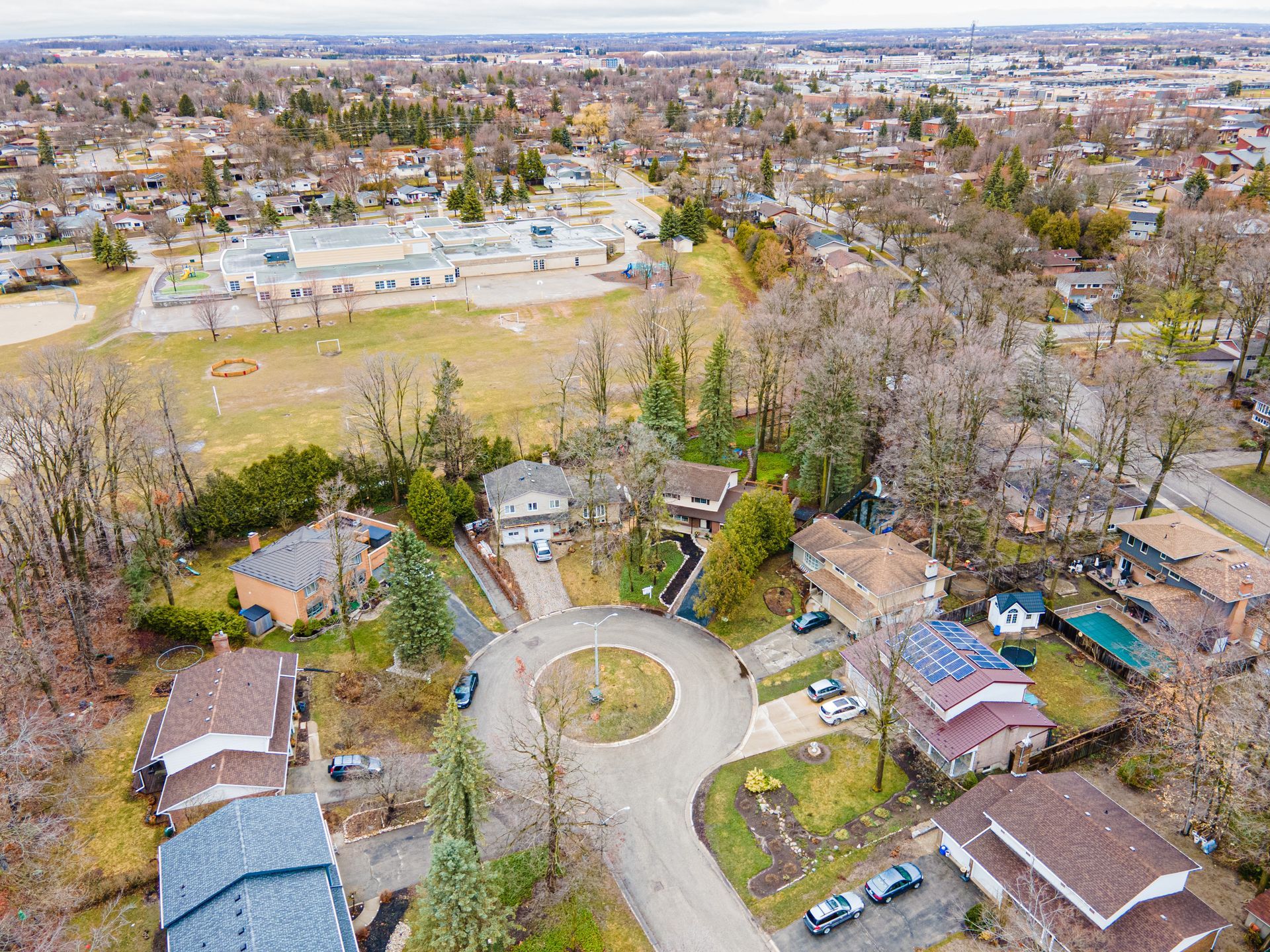 Aerial view of suburban houses on a cul-de-sac near a schoolyard with bare trees and overcast sky.