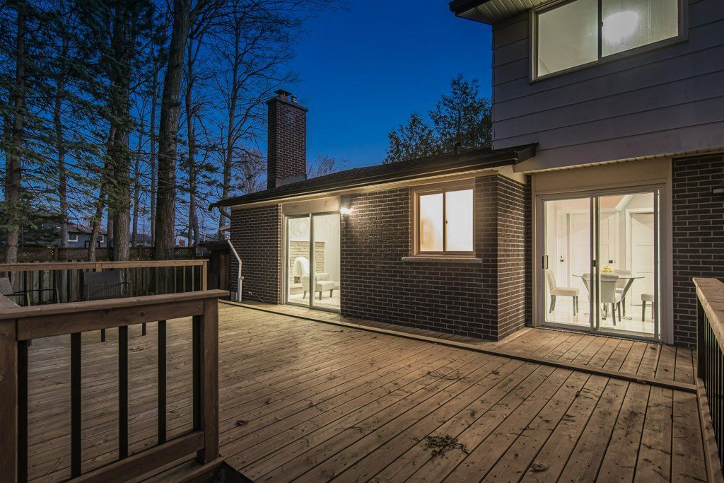 Wooden deck at dusk; brick building with lit windows, chimney, surrounded by trees.