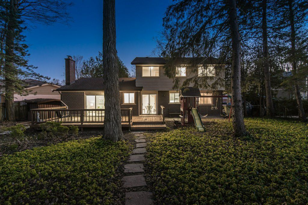 Backyard view of a two-story house at dusk, with lights on. A stone path leads to a wooden deck.
