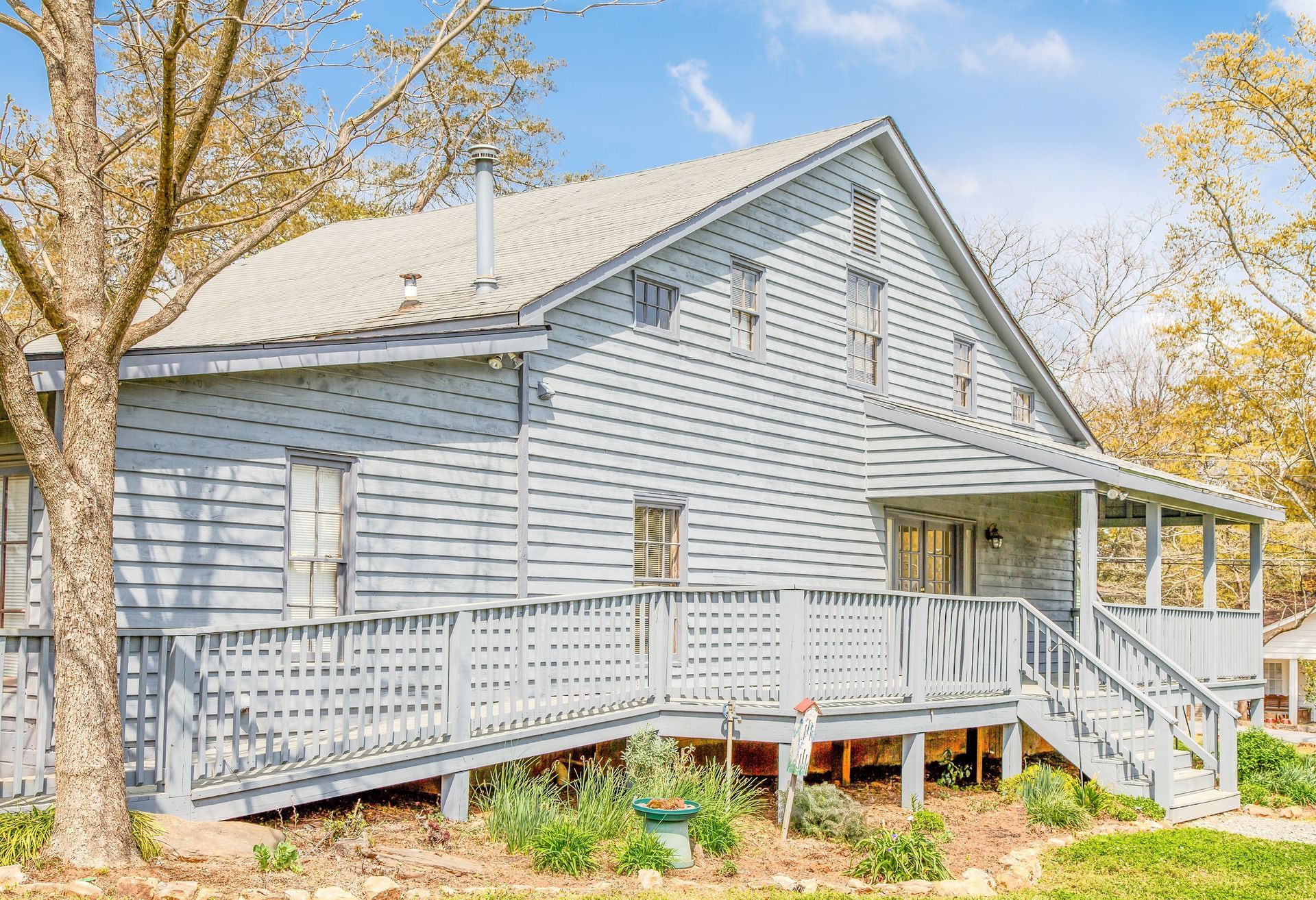 Light blue two-story house with a wooden deck, a ramp, and porch. Surrounding trees under a blue sky.