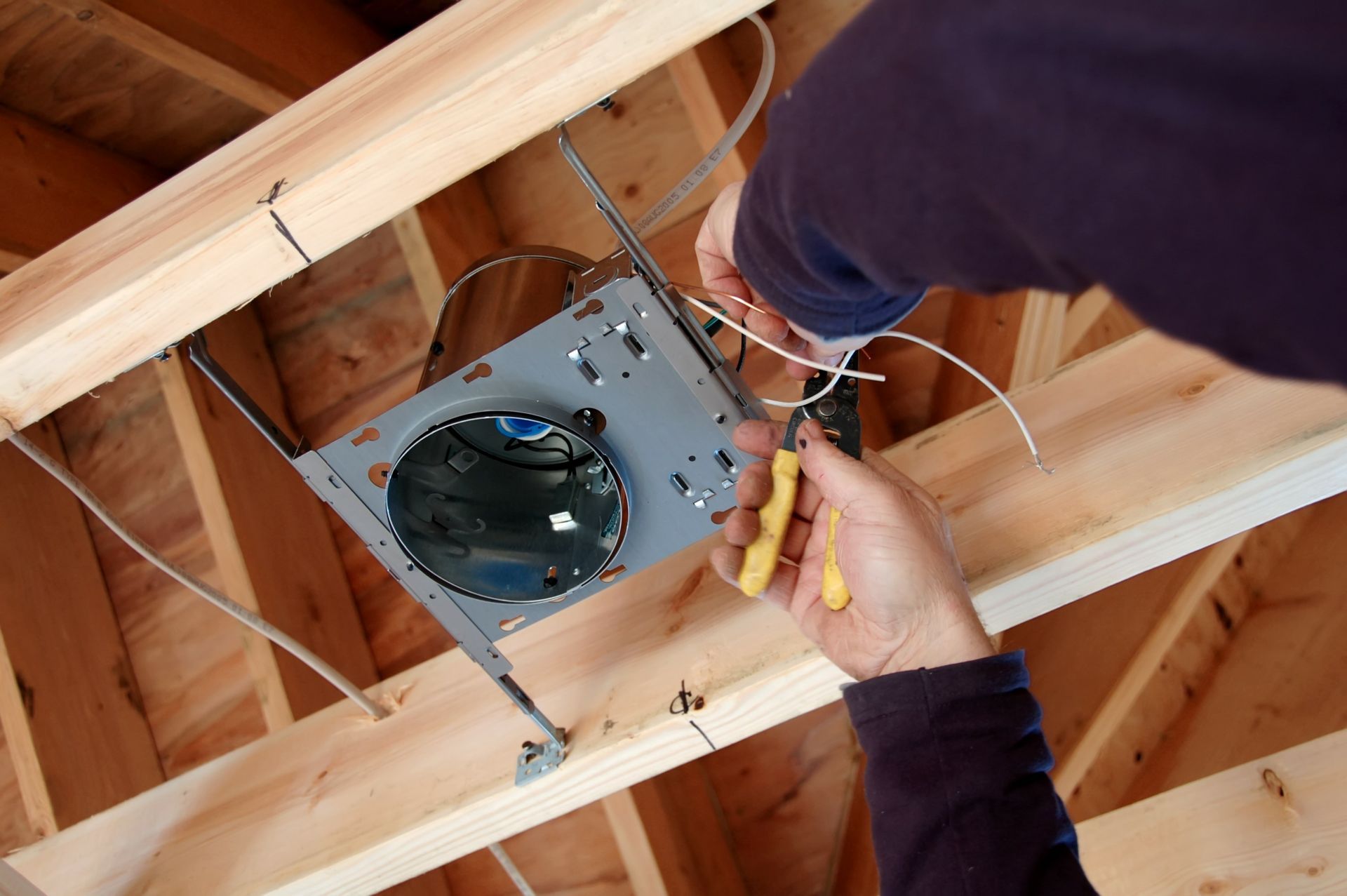 Electrician installing recessed light fixture in a wooden ceiling, using wire strippers.