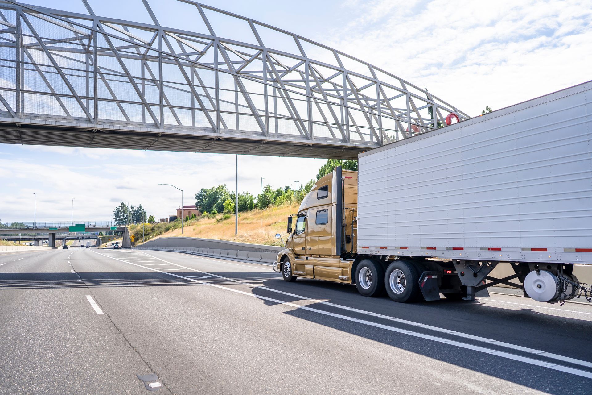 A semi truck is driving under a bridge on a highway.