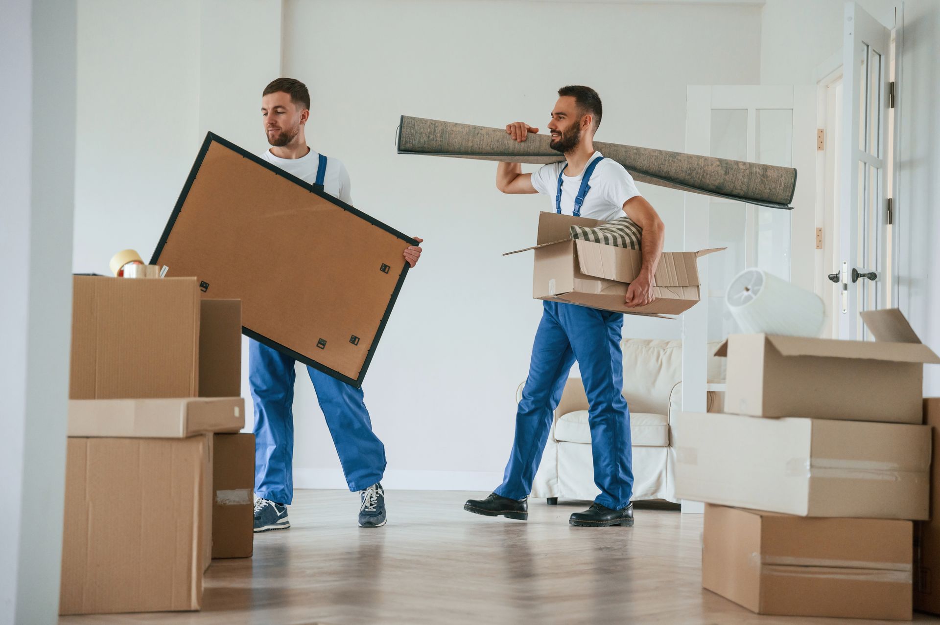 Two men are carrying boxes and a rug in a living room.