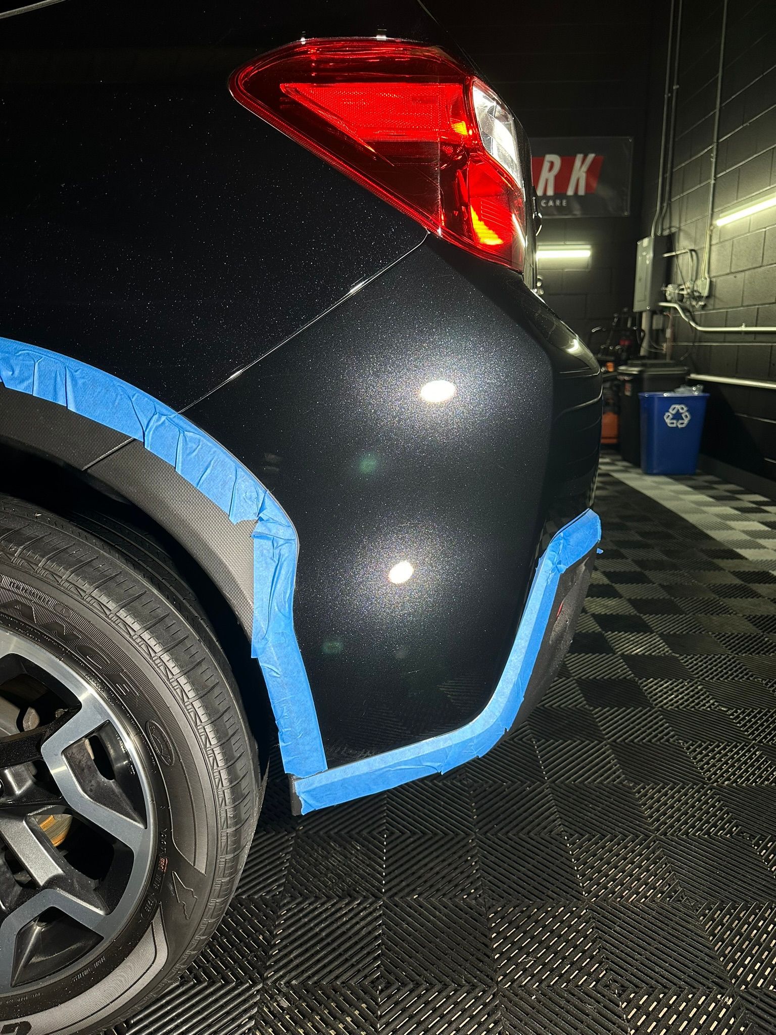 A black car with blue tape on the fender is in a garage.