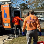 Two plumbers in orange shirts work beside a service van on a suburban lawn.