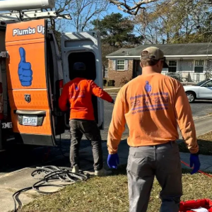 Two plumbers in orange shirts work beside a service van on a suburban lawn.