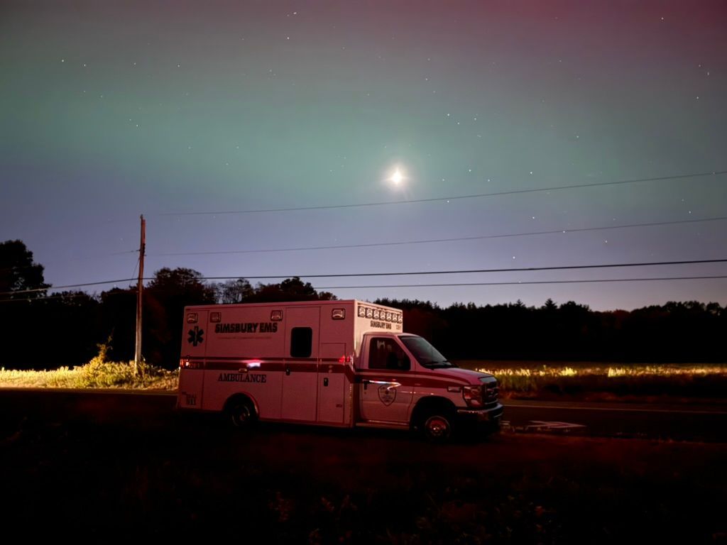 A white and red Simsbury ambulance is parked on the side of the road with the aurora borealis visible.