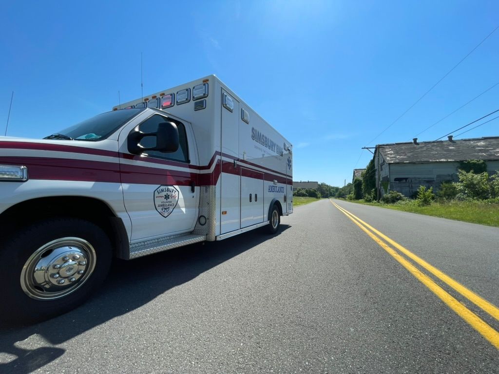 A white and red Simsbury ambulance parked on the side of the road near a tobacco barn.