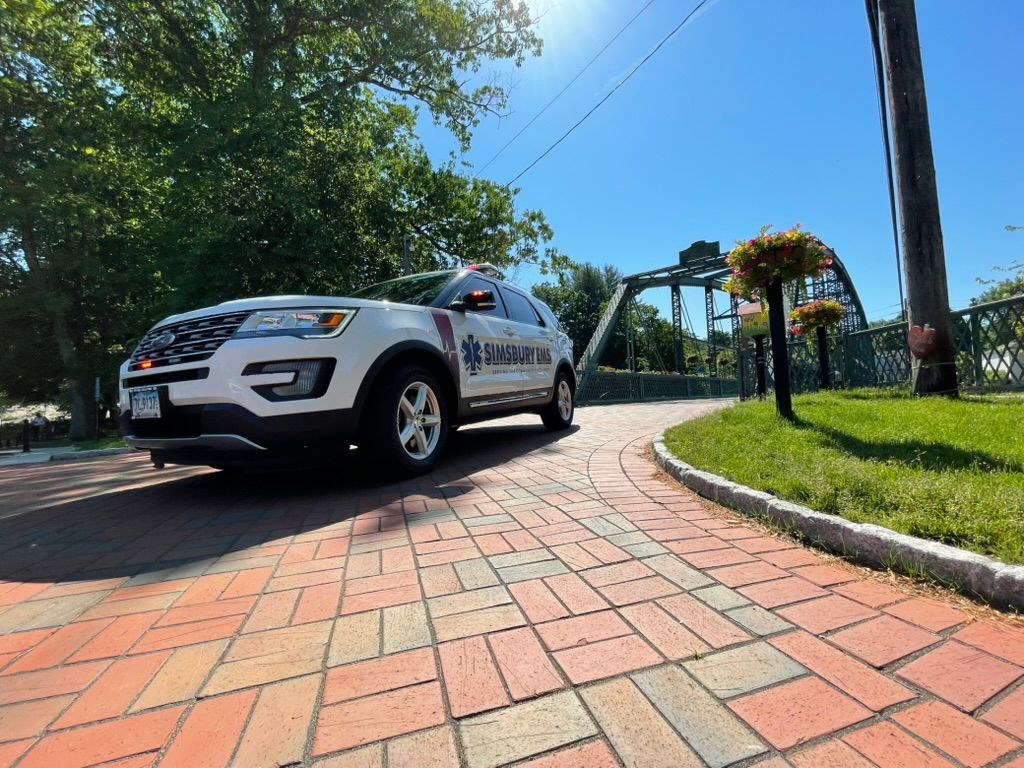 A white ambulance flycar is parked on a brick sidewalk next to a flower bridge in Simsbury, CT.