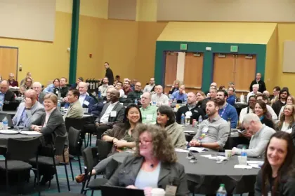 Audience seated at round tables in a conference room, many looking toward the front and smiling.