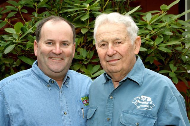 Two men are posing for a picture together in front of a bush.