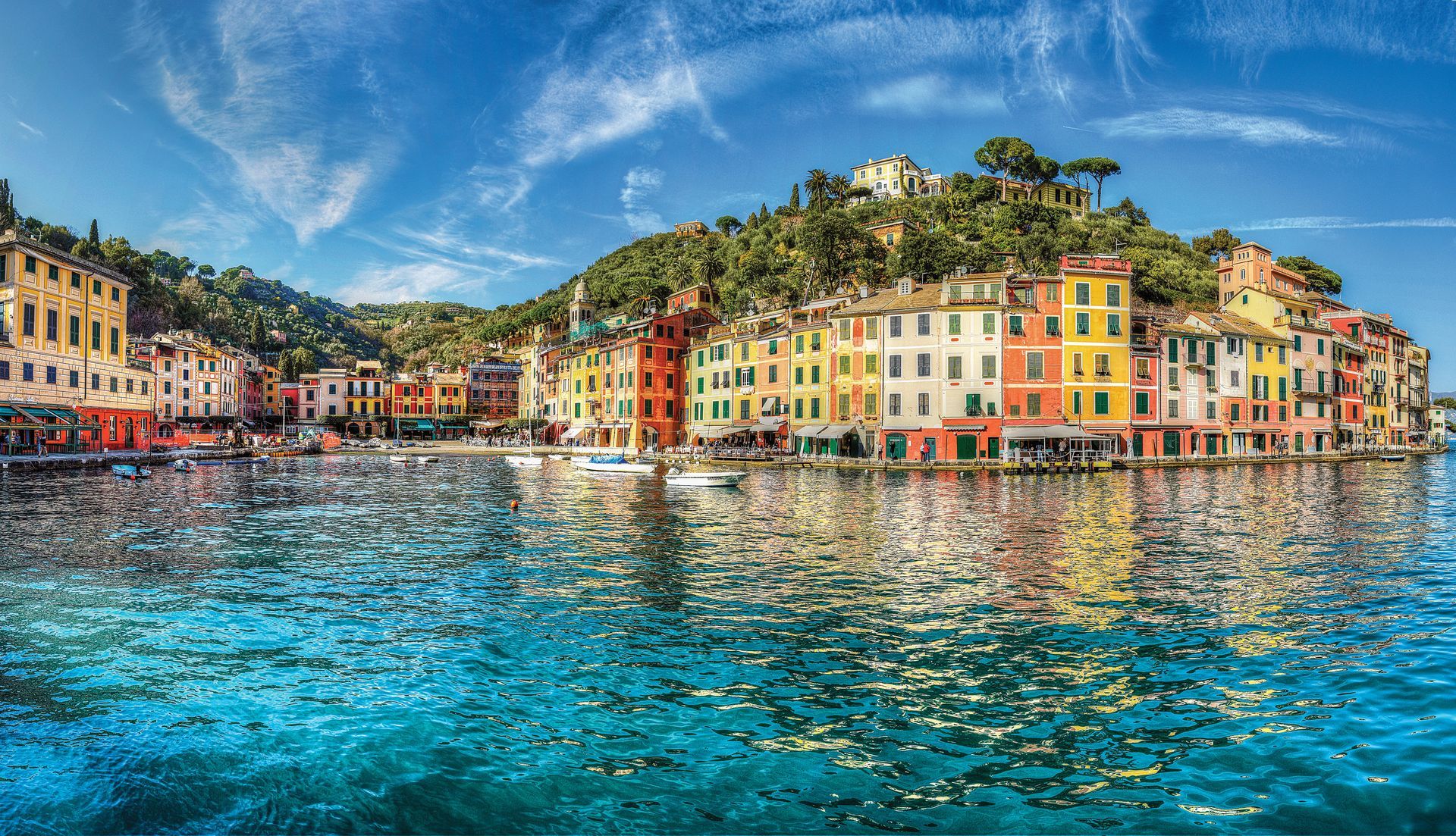 An aerial view of a large cruise ship docked at a paved pier next to a coastal town with red-roofed buildings.