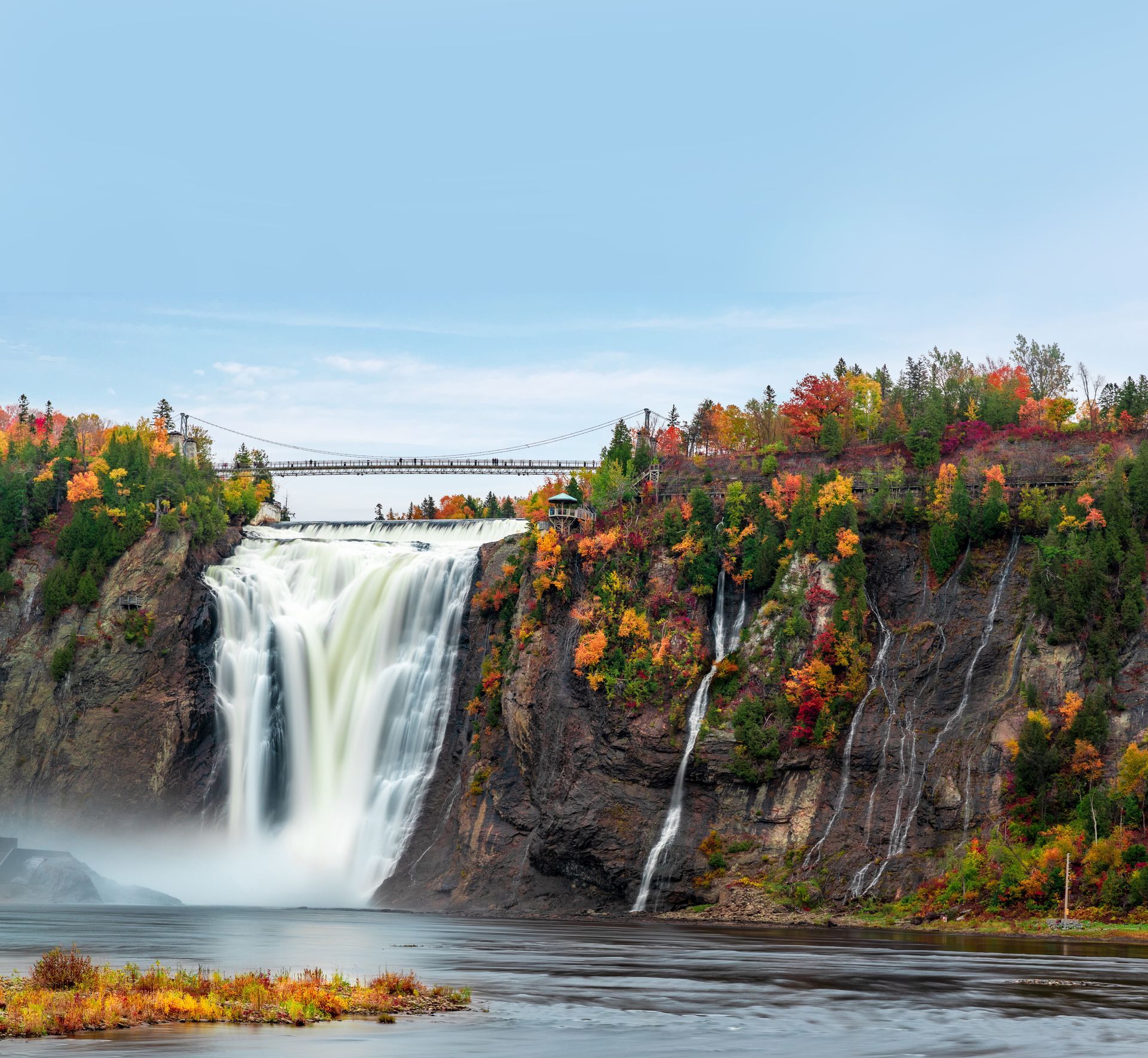 A waterfall is surrounded by trees and a bridge in the background.