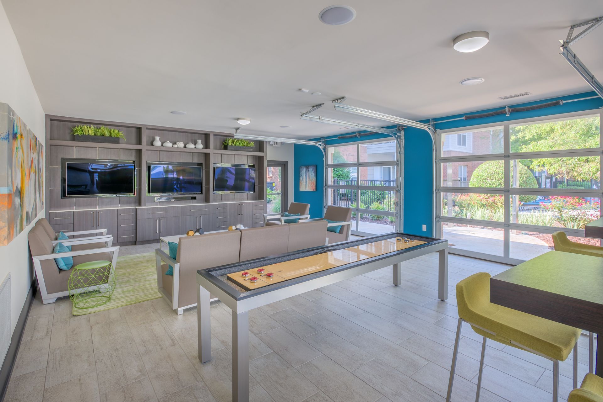 A modern lounge area with a shuffleboard table, multiple TVs, and large windows. Light wood floors and teal accents.