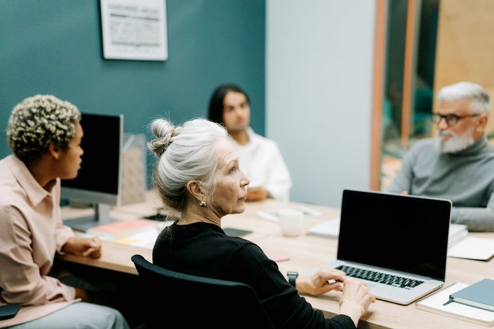 People in a meeting, woman with gray hair uses laptop, others listen, in a brightly lit room.