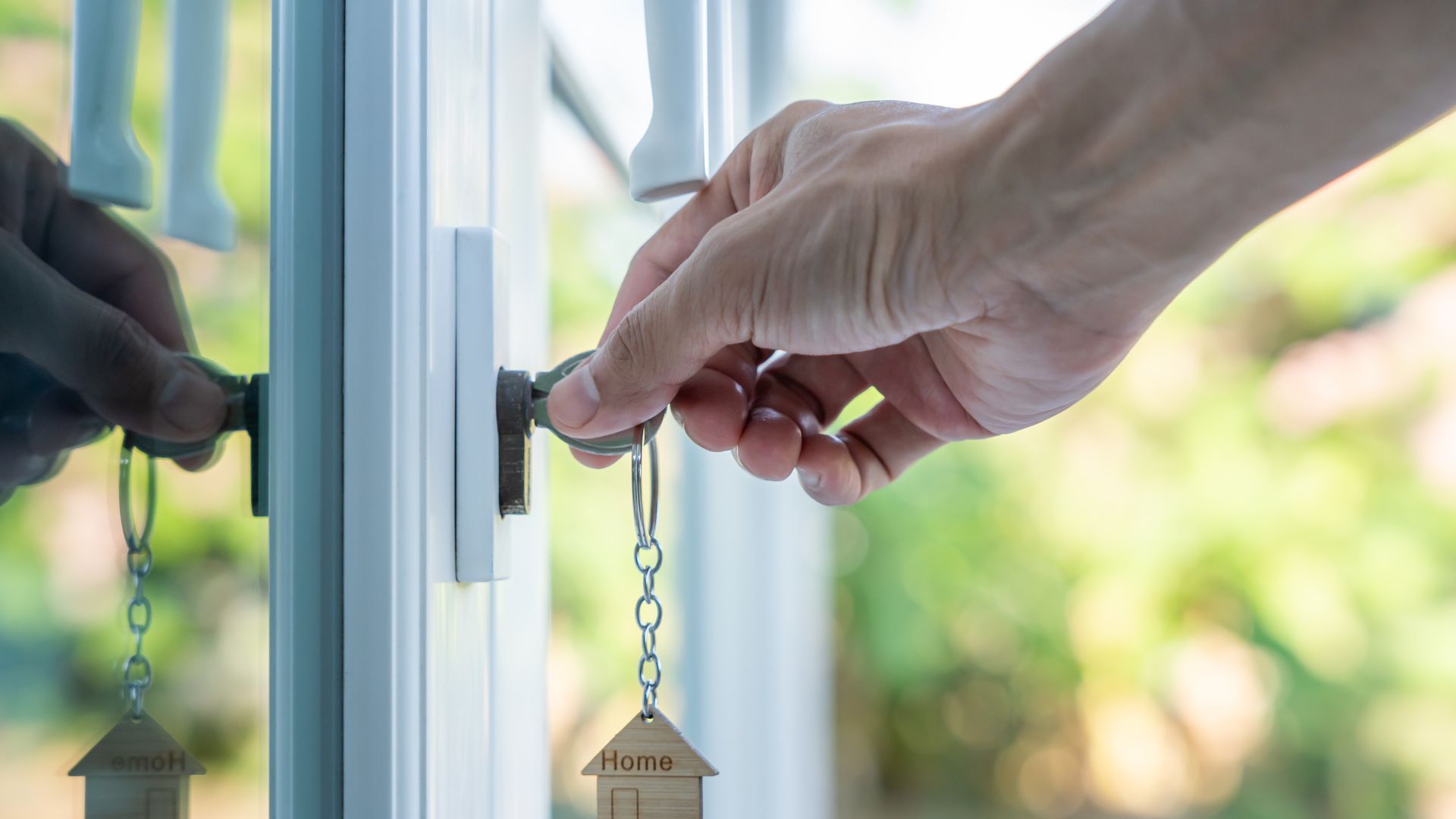 Hand turning a key in a door lock, with a house-shaped keychain, blurred green background.
