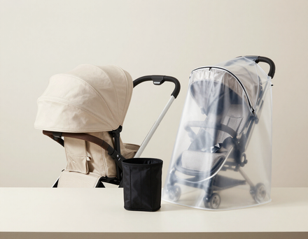Sobre una mesa, hay un cochecito de bebé de color beige, un pequeño portavasos negro y un segundo cochecito cubierto con una lona transparente para la lluvia.