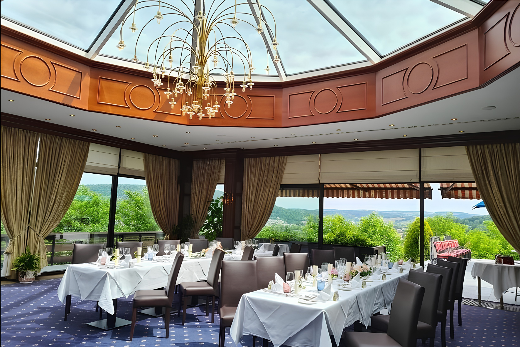Salle à manger élégante avec plafond vitré, lustre, longues tables et vue sur collines verdoyantes et arbres.