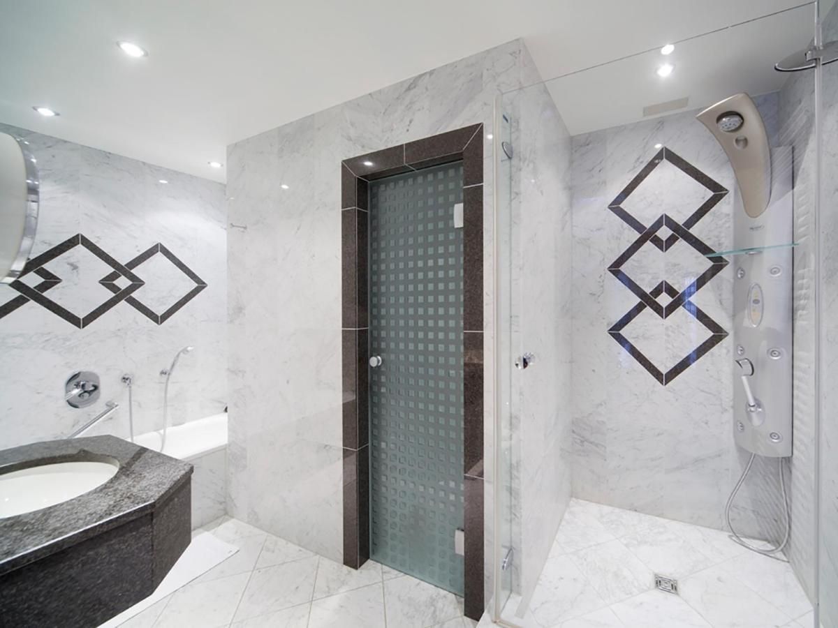 Salle de bain luxueuse en marbre avec porte vitrée, pommeau de douche et accents de carrelage géométrique noir et blanc.