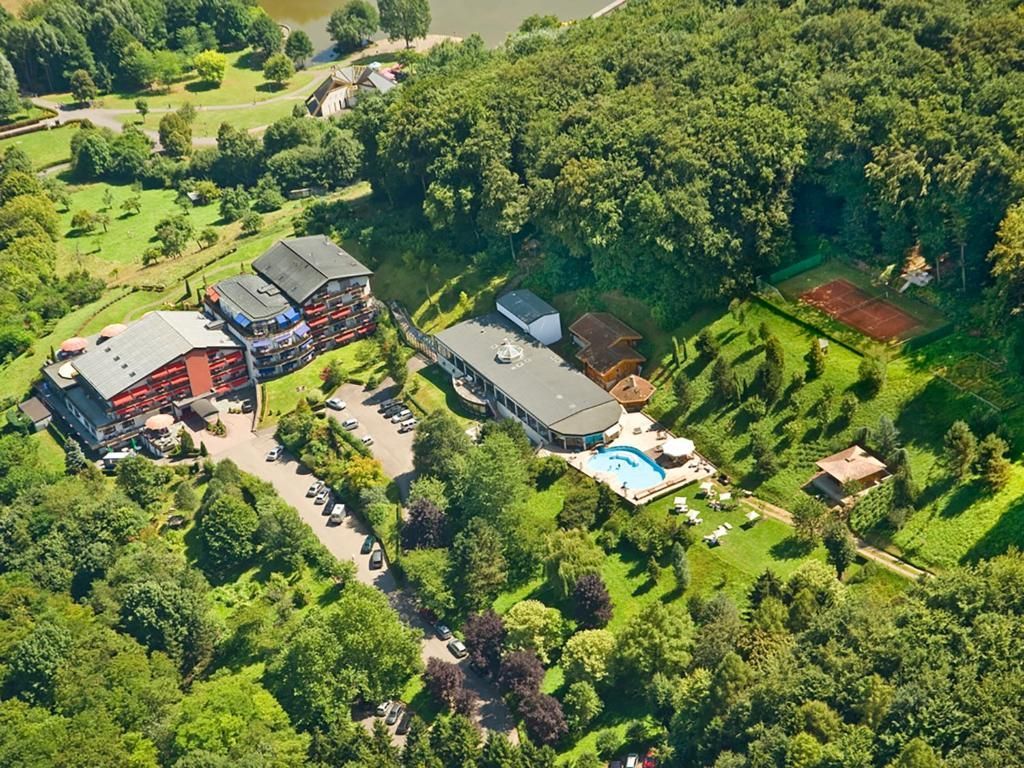 Vue aérienne d'un complexe avec plusieurs bâtiments, une piscine, un court de tennis et un parking entouré d'arbres et de feuillages verdoyants.