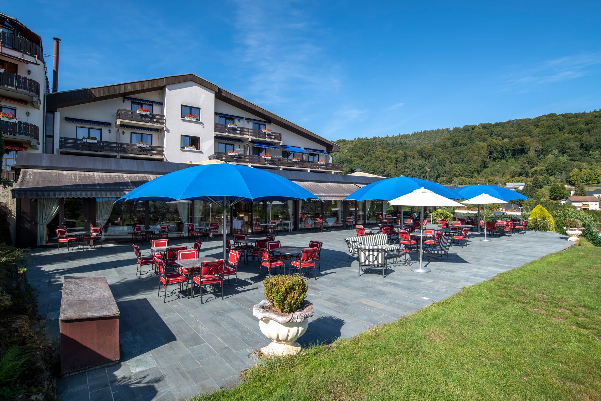 Patio de l'hôtel avec tables, chaises et parasols bleus, donnant sur une colline.
