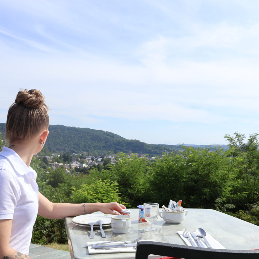 Une personne est assise à une table en terrasse, profitant d'une vue pittoresque sur une colline boisée et une petite ville.