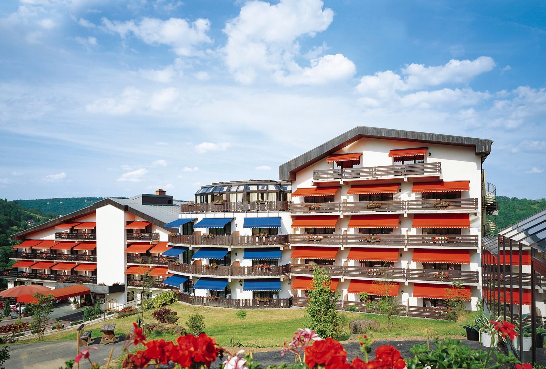 Extérieur de l'hôtel avec plusieurs étages, auvents rouges et bleus et balcons, entouré d'herbe verte et de roses rouges.