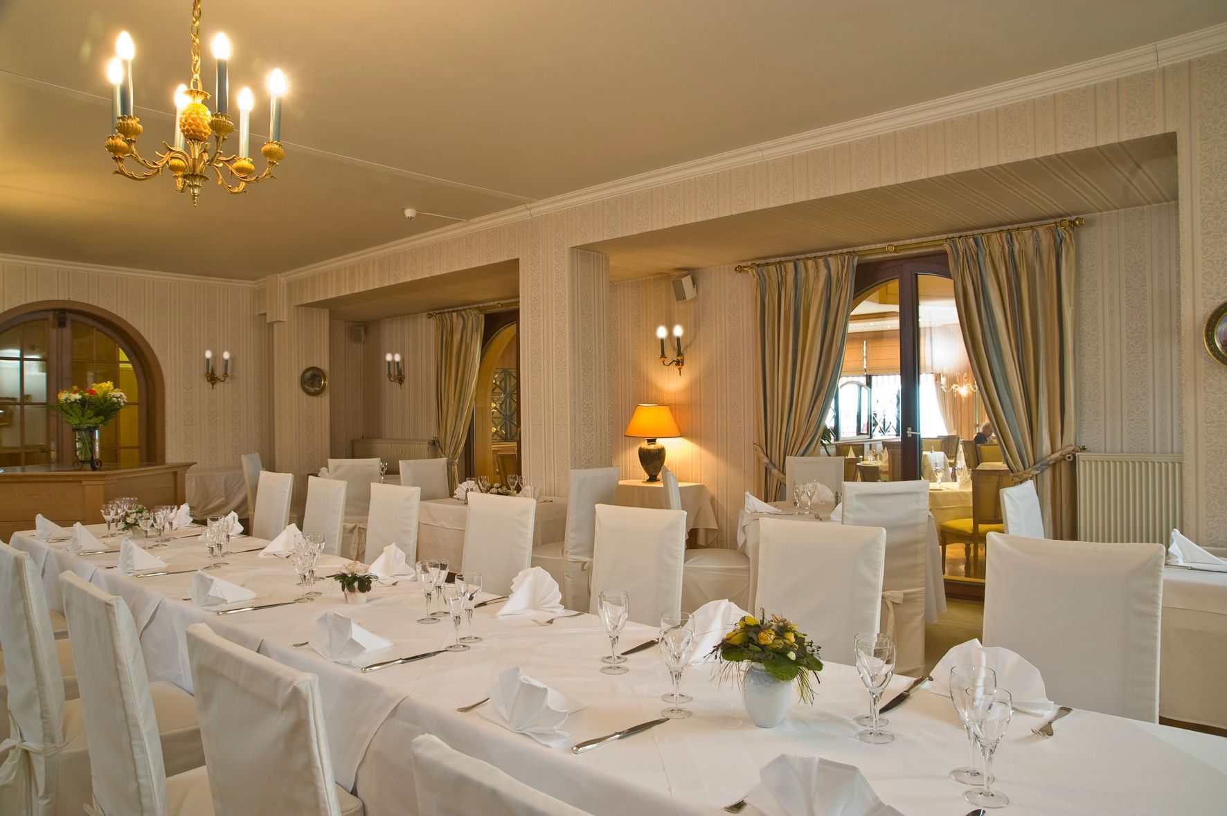 Salle à manger élégante avec longues tables nappées de blanc, chaises blanches et lustre doré au plafond.