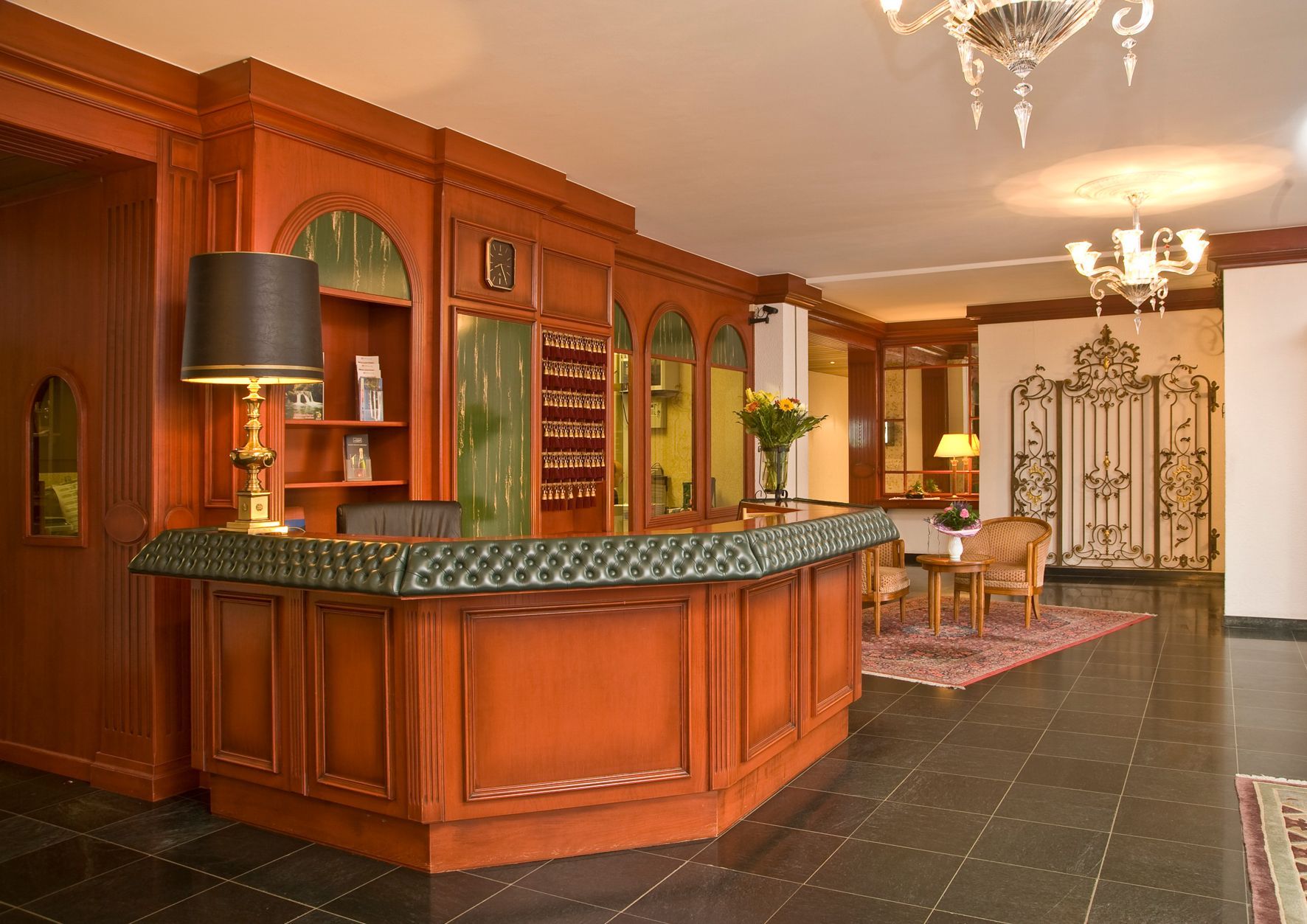 Comptoir d’hôtel en bois avec lampe noire, dans un hall élégant avec sol en marbre et grille en fer forgé.