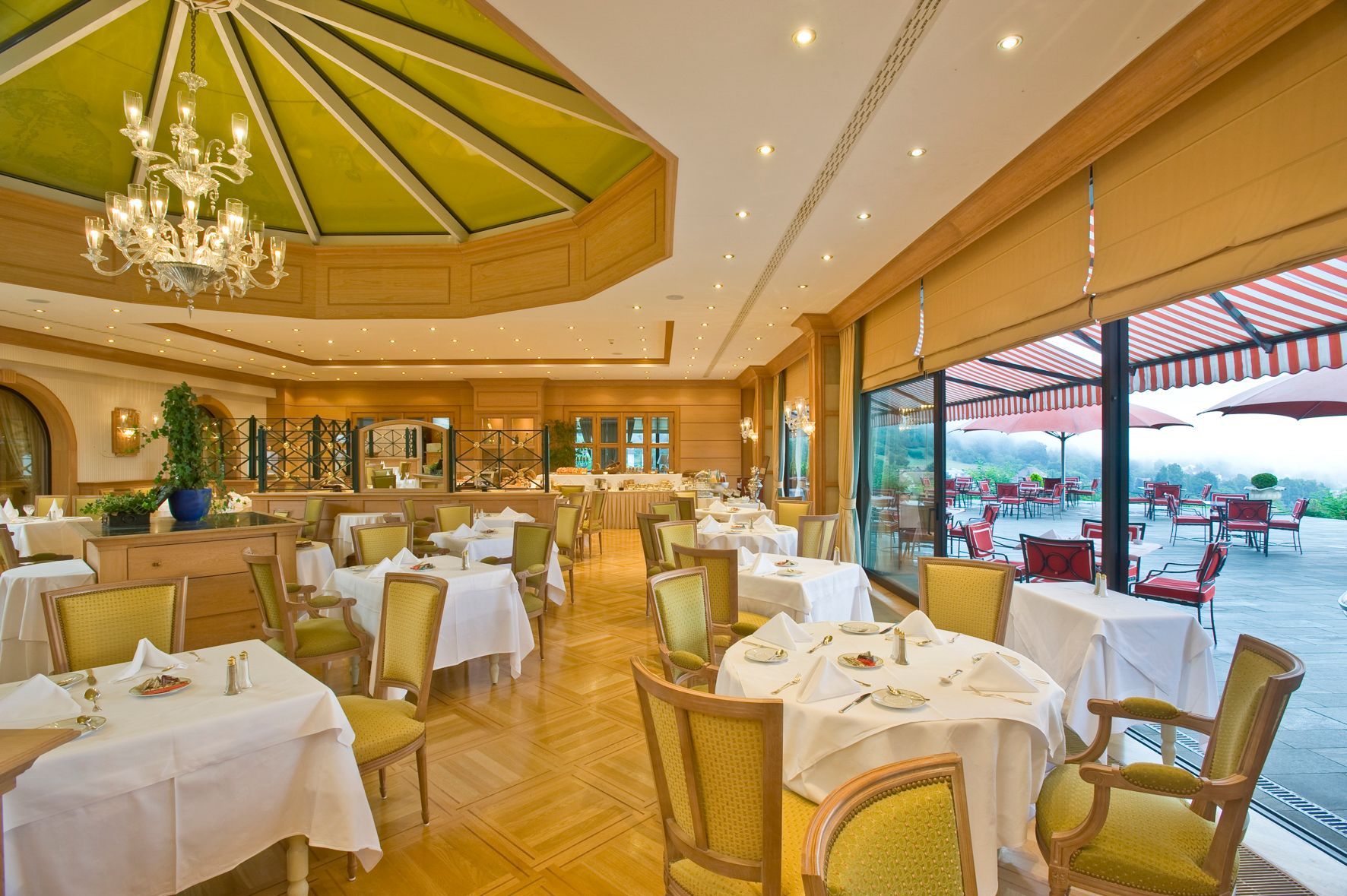 Intérieur de restaurant avec tables blanches, chaises vertes, lustre et vue sur terrasse extérieure.