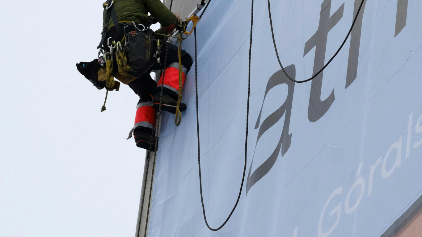 Person rappelling down a blue sign. They wear safety gear, including a harness and red leg protectors.