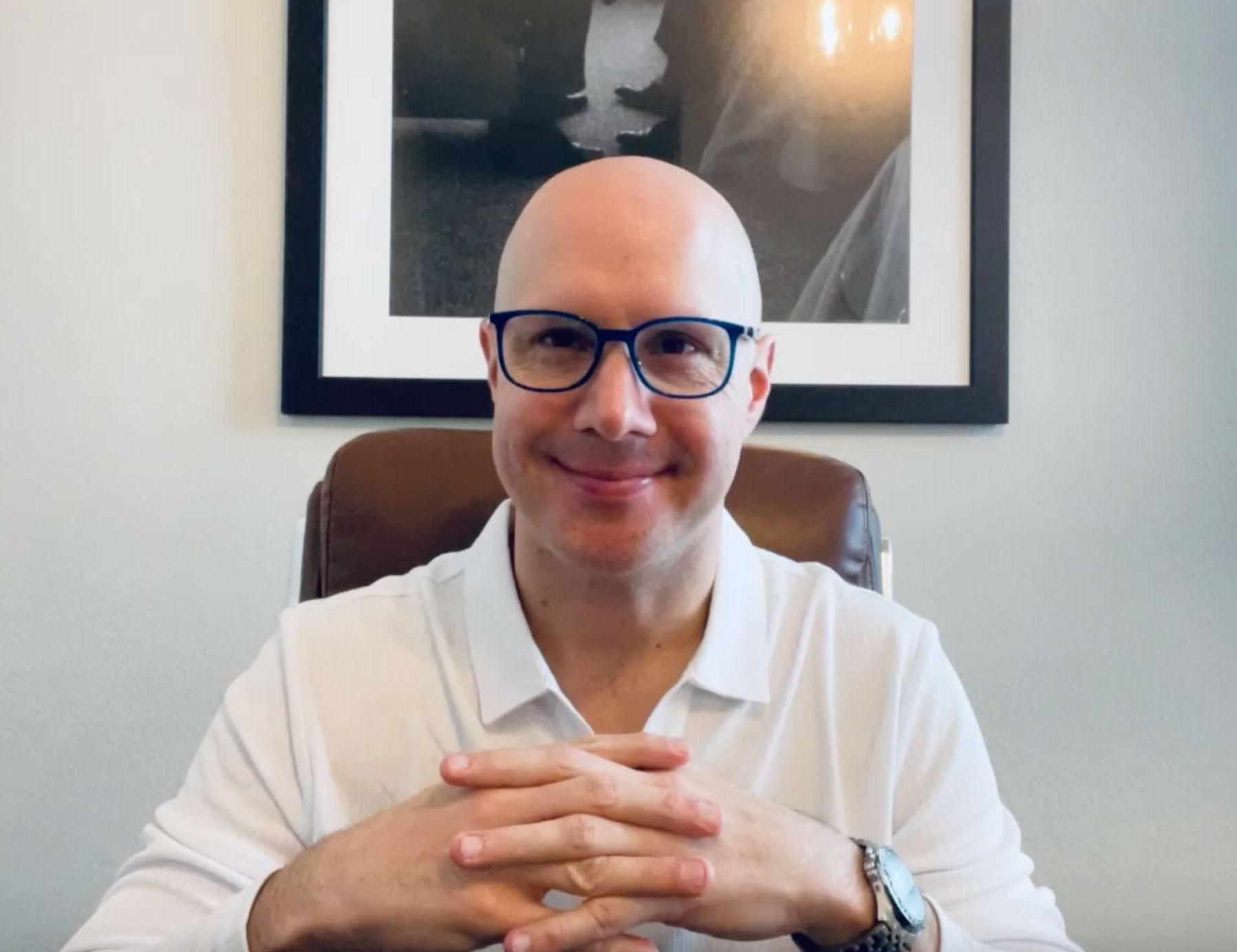 A bald man wearing glasses is sitting in a chair with his hands folded.