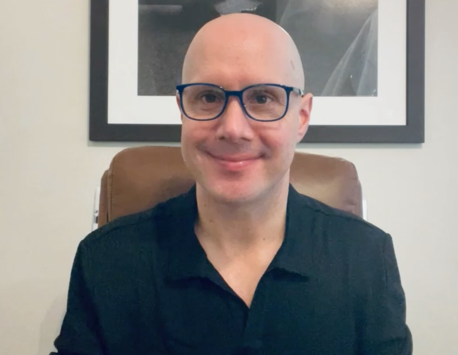 A bald man wearing glasses and a black shirt is sitting in a chair.