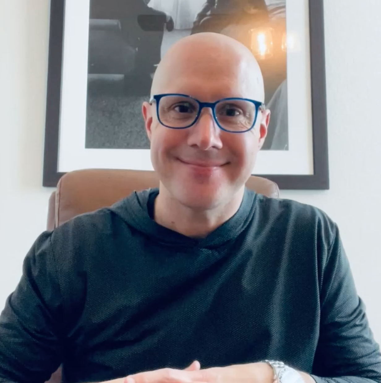 A bald man wearing glasses is sitting in front of a framed picture