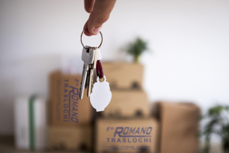 Mano che tiene un mazzo di chiavi con un'etichetta bianca, le caselle sullo sfondo suggeriscono un trasloco.