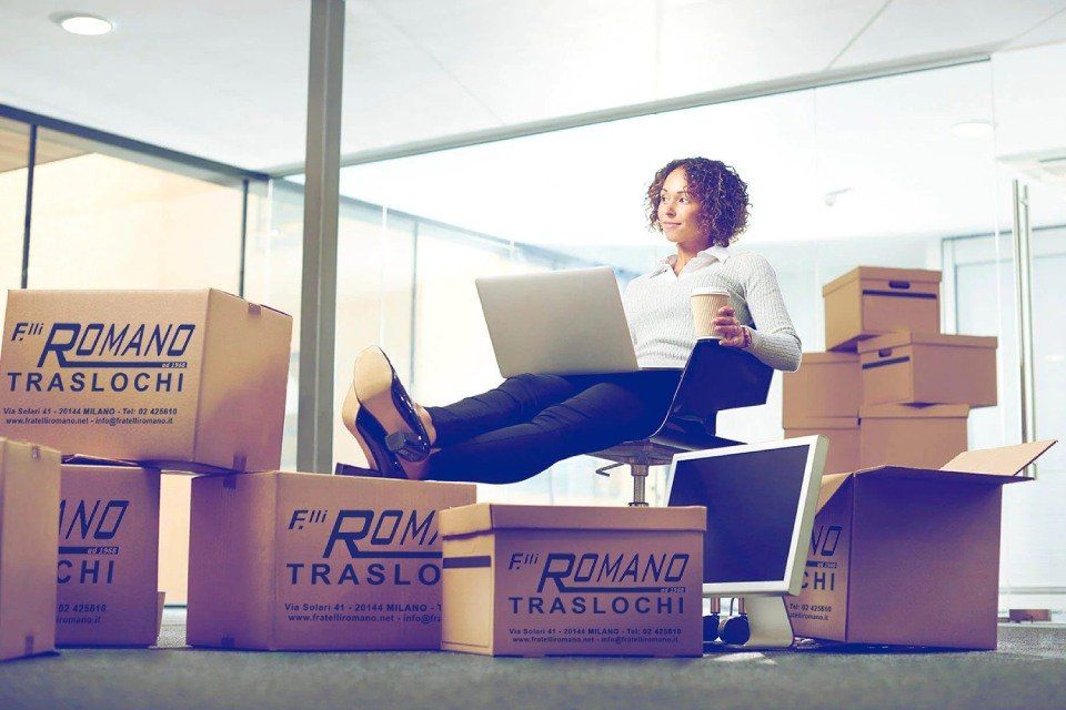 Donna seduta su una sedia da ufficio con i piedi sollevati, che usa un computer portatile, circondata da scatoloni da trasloco. Tiene in mano una tazza di caffè e sembra rilassata.
