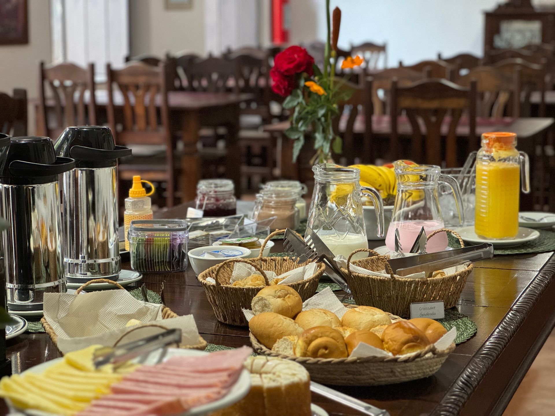 Buffet de café da manhã com pães, frutas, sucos e café em uma mesa de madeira em uma sala de jantar.