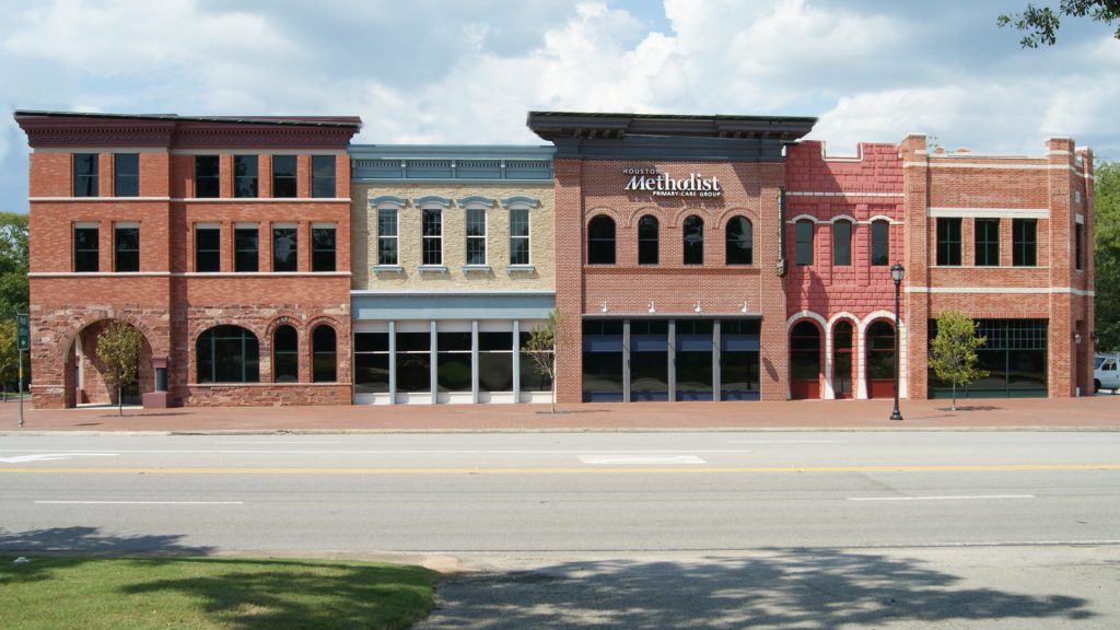Row of brick storefronts on a sunny day. The buildings have varied designs and colors, with large windows and awnings.
