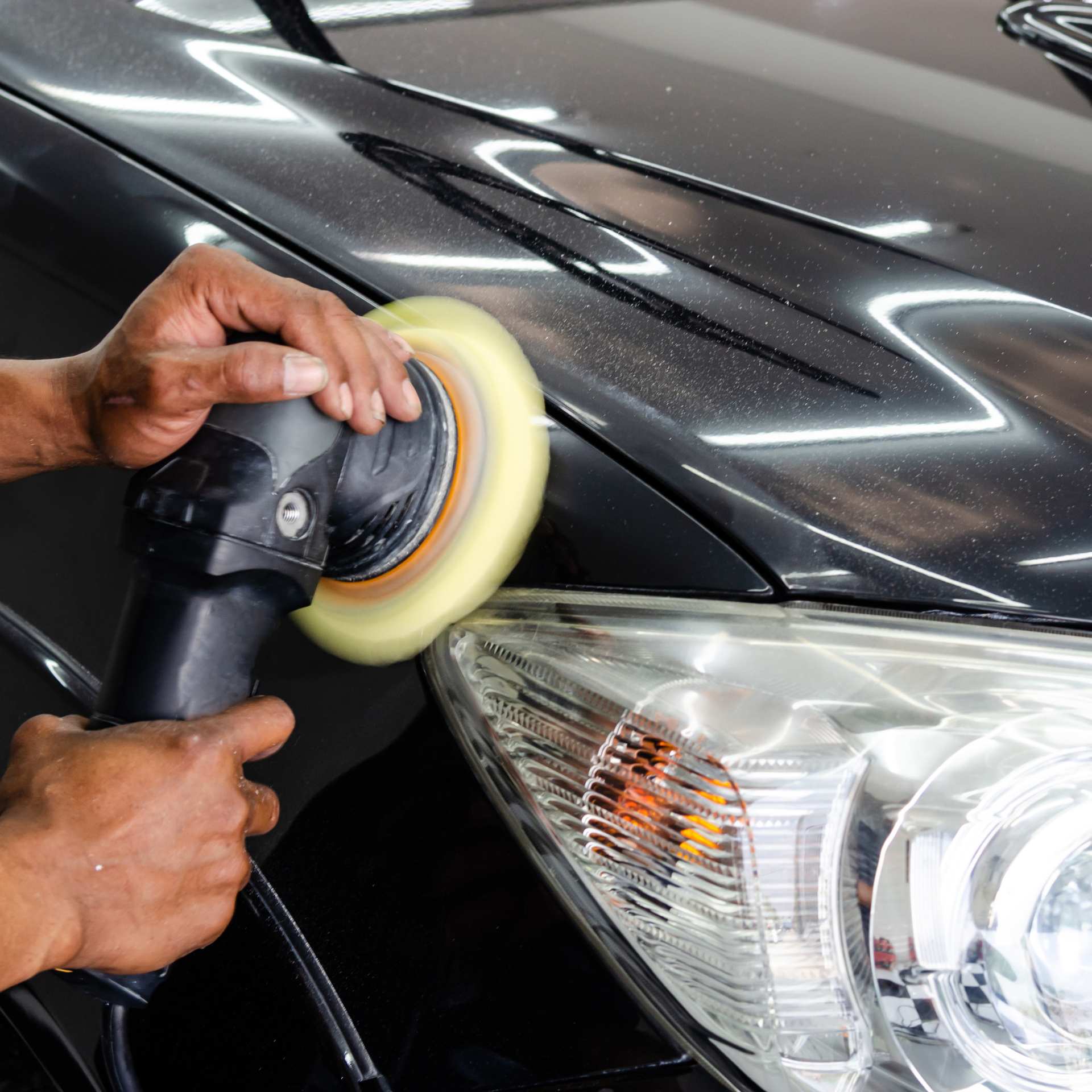 Person using a buffer to polish a black car's hood, near a headlight.