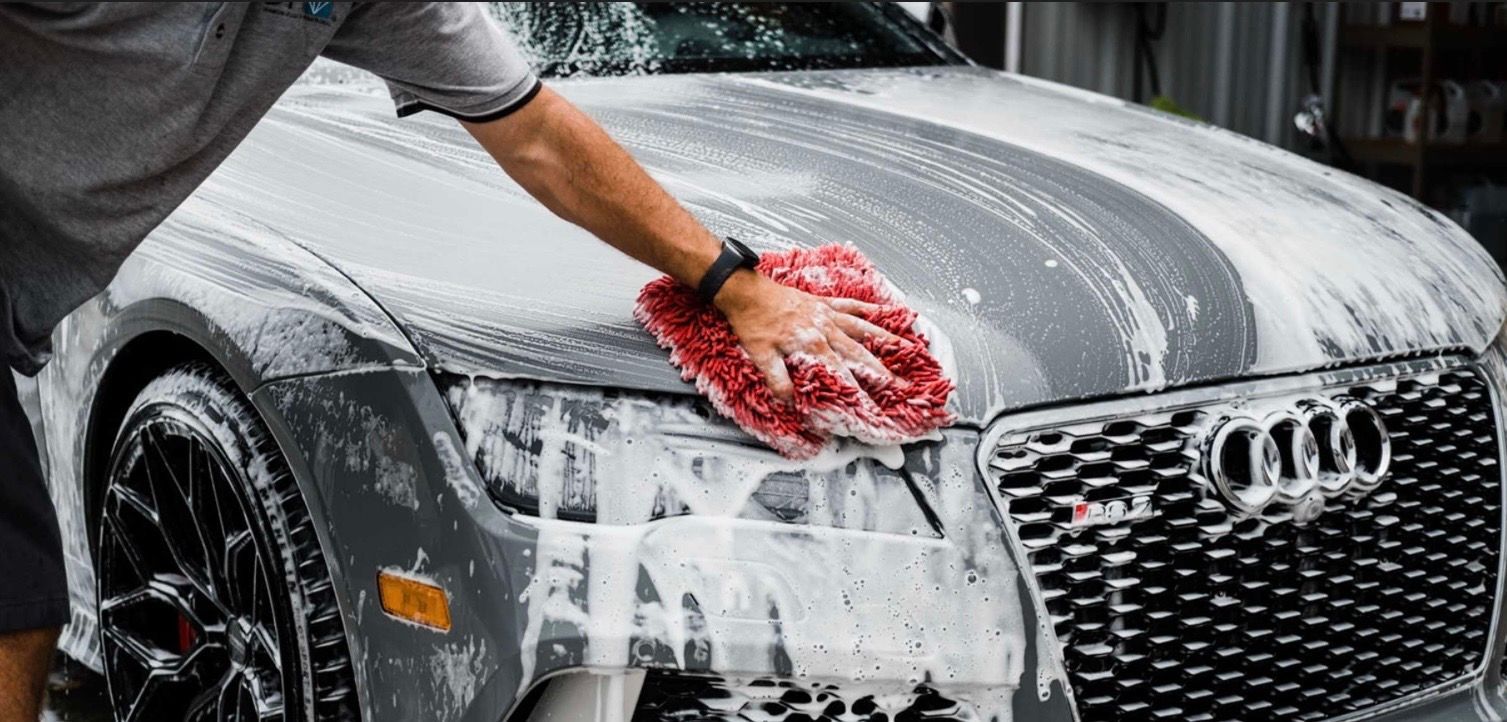 A person washing a gray Audi with a red wash mitt covered in soapy suds.