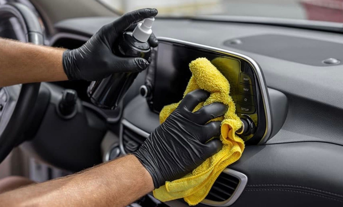 Person wearing black gloves cleaning a car dashboard with a spray bottle and yellow cloth.