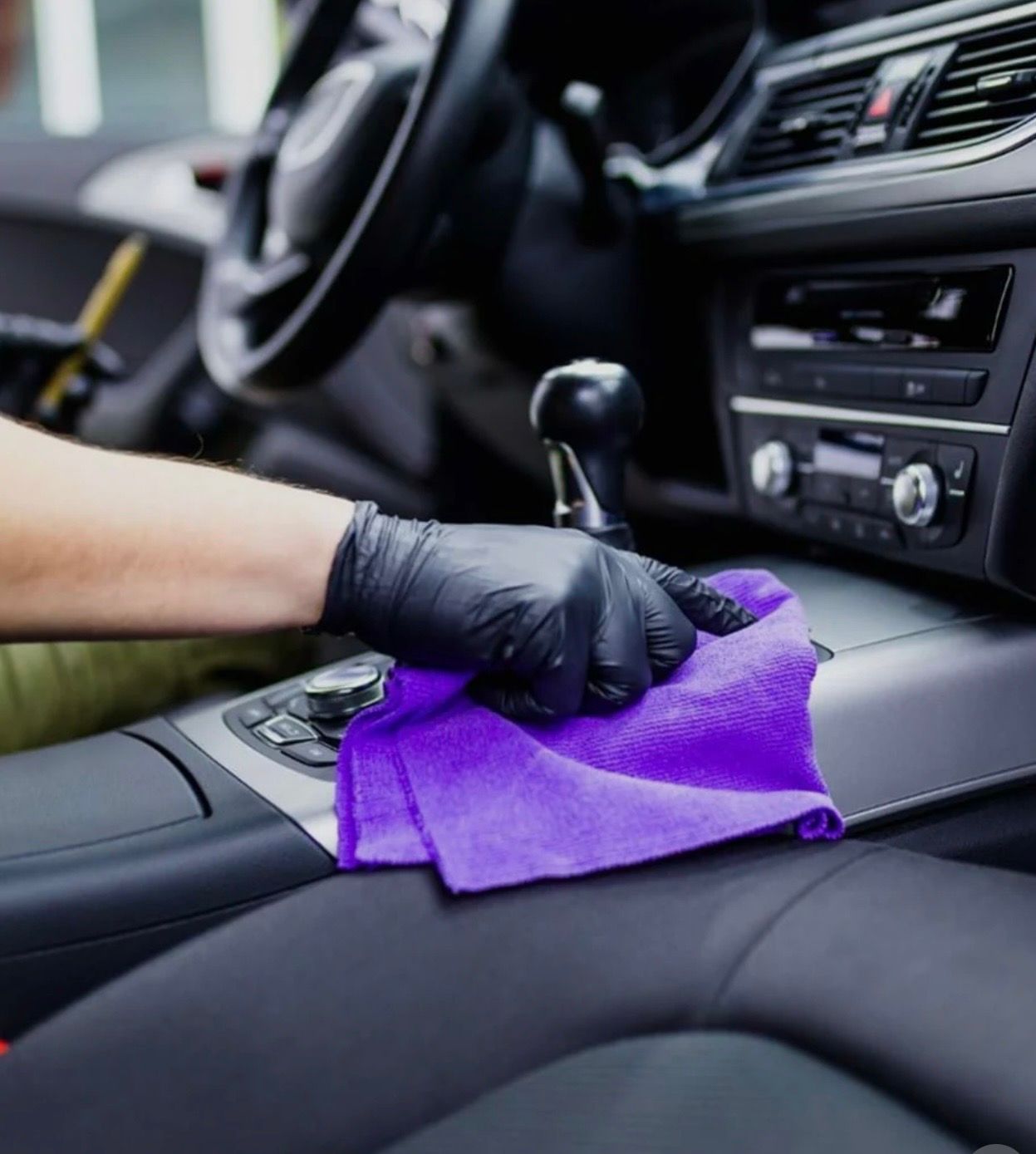 Gloved hand wiping a purple cloth on a car's interior console. Black and silver dashboard details visible.