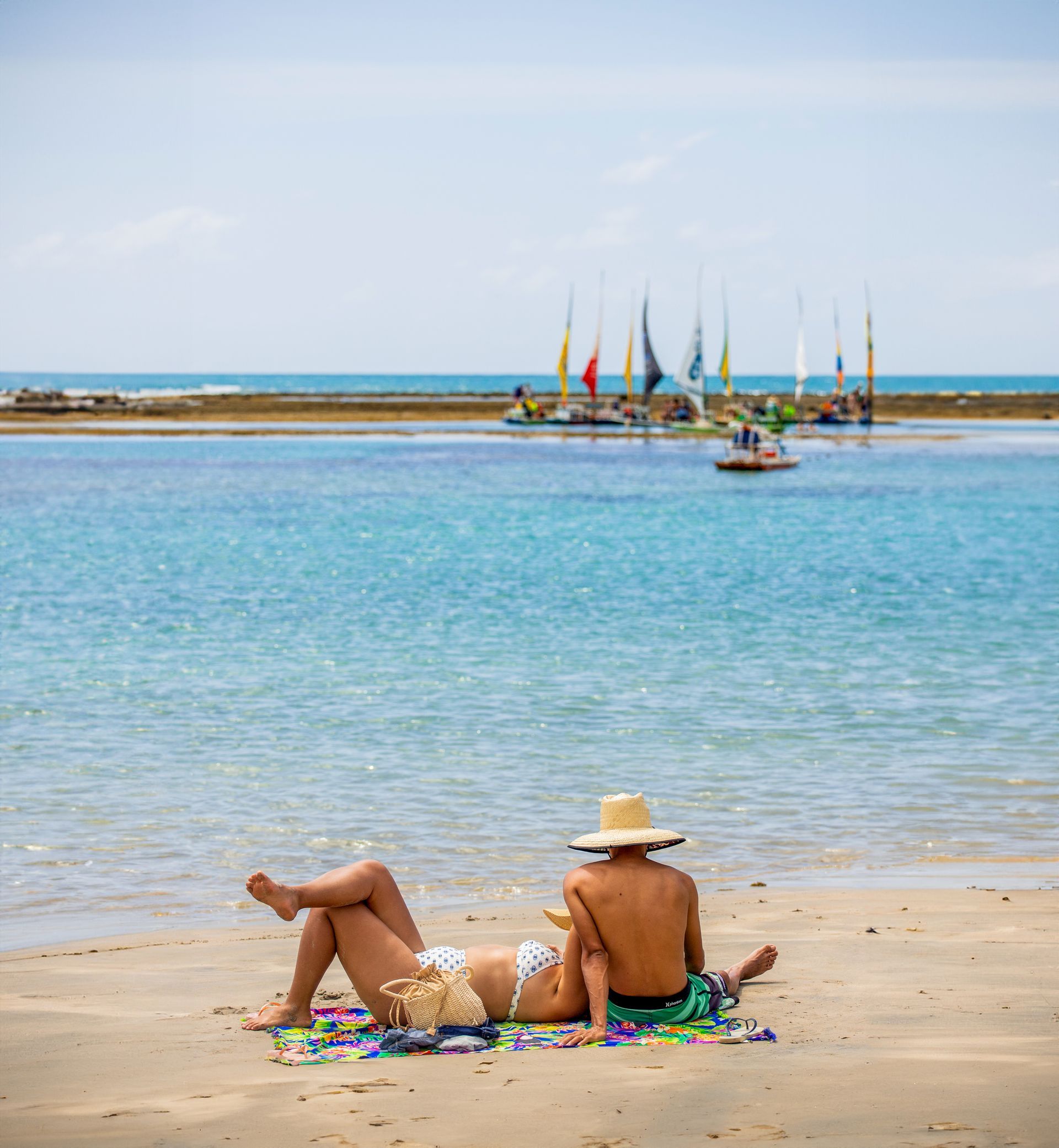 Casal relaxando na praia, oceano com veleiros ao fundo.