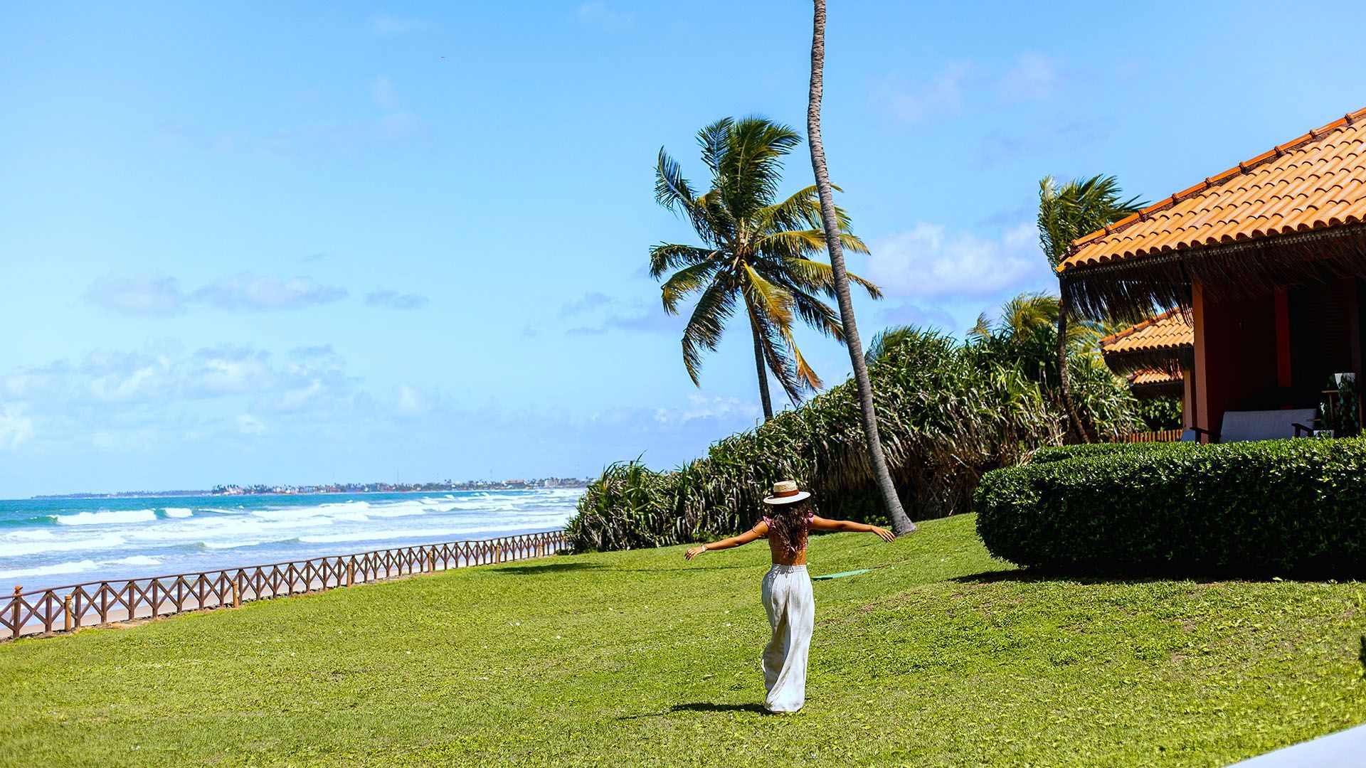 Piscina em um resort com espreguiçadeiras, palmeiras e o oceano sob um céu azul claro.