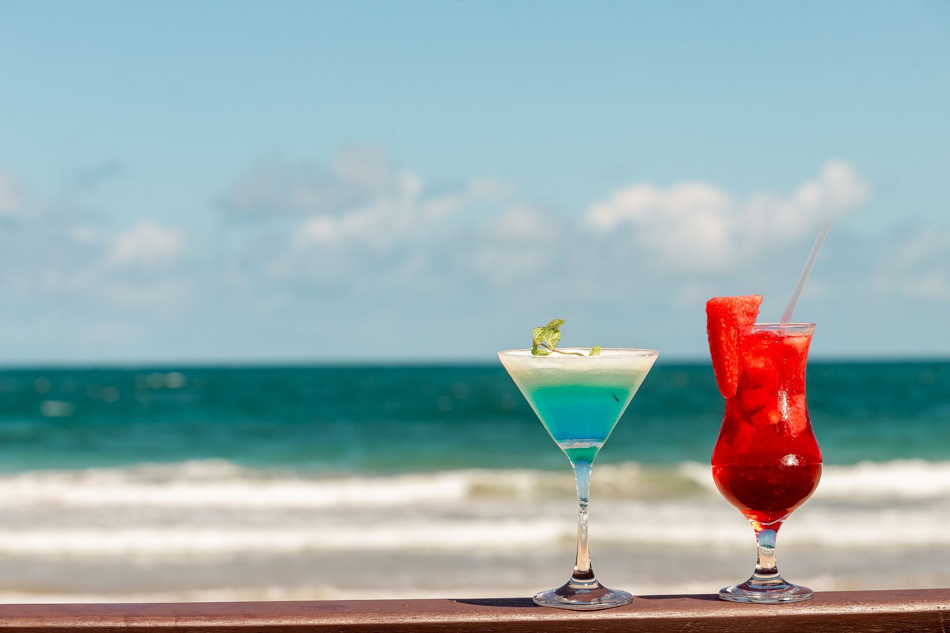 Dois coquetéis em uma superfície de madeira, com o oceano e o céu ao fundo.