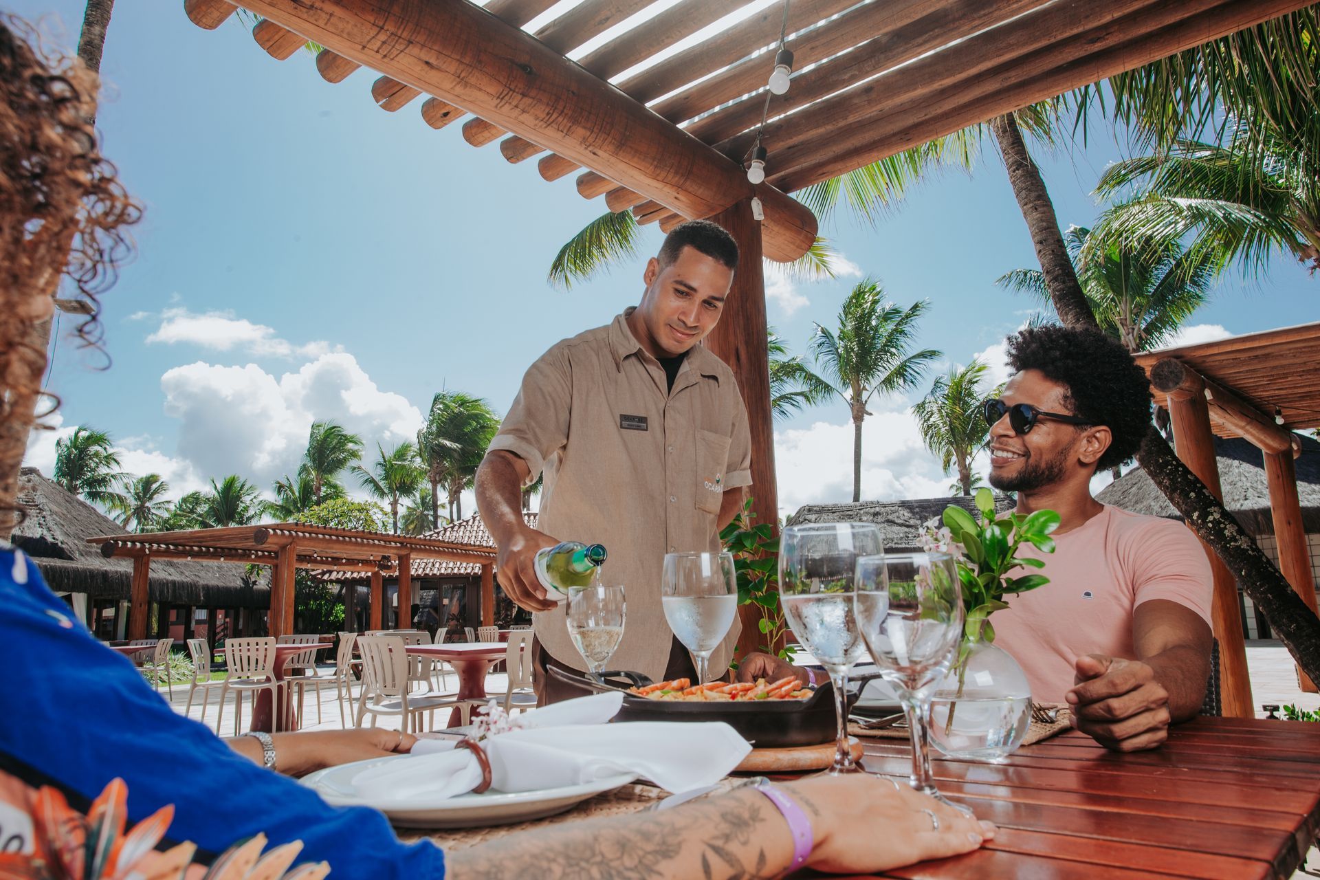 Garçom serve bebida para casal em mesa ao ar livre, cenário de praia.