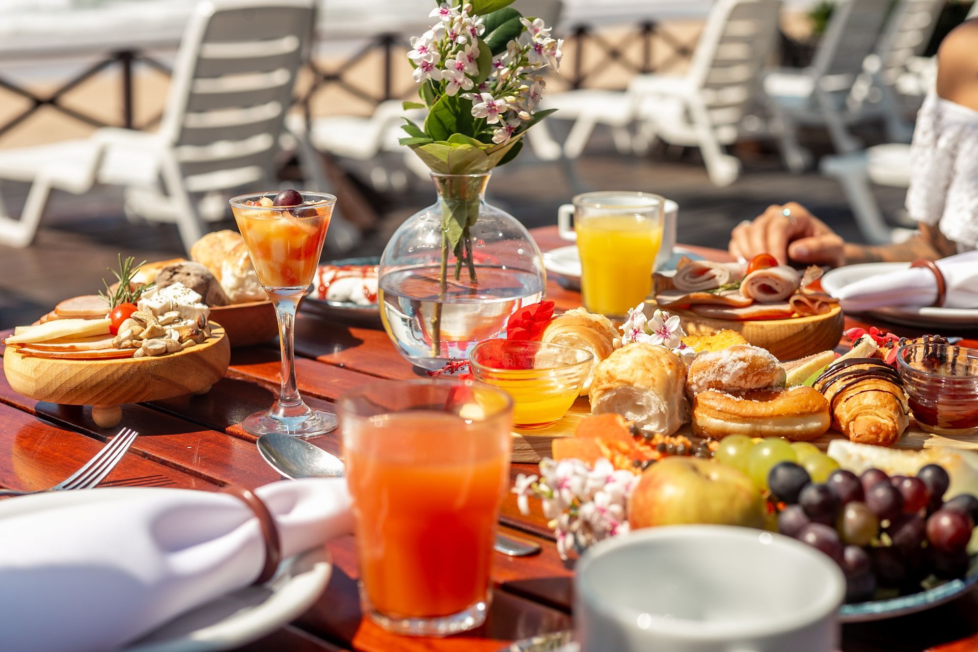 Café da manhã: pão, frutas, suco, frios e flores sobre uma mesa de madeira, ao ar livre, com espreguiçadeiras ao fundo.