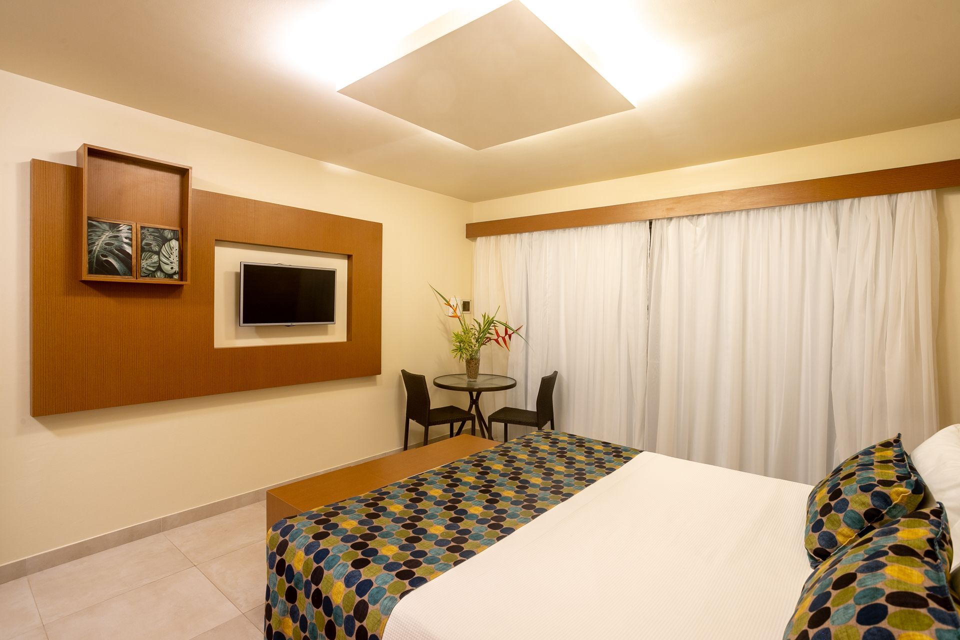 Quarto de hotel com cama, TV, mesa e cadeiras. Detalhes em madeira, cortinas brancas e uma luminária retangular no teto.