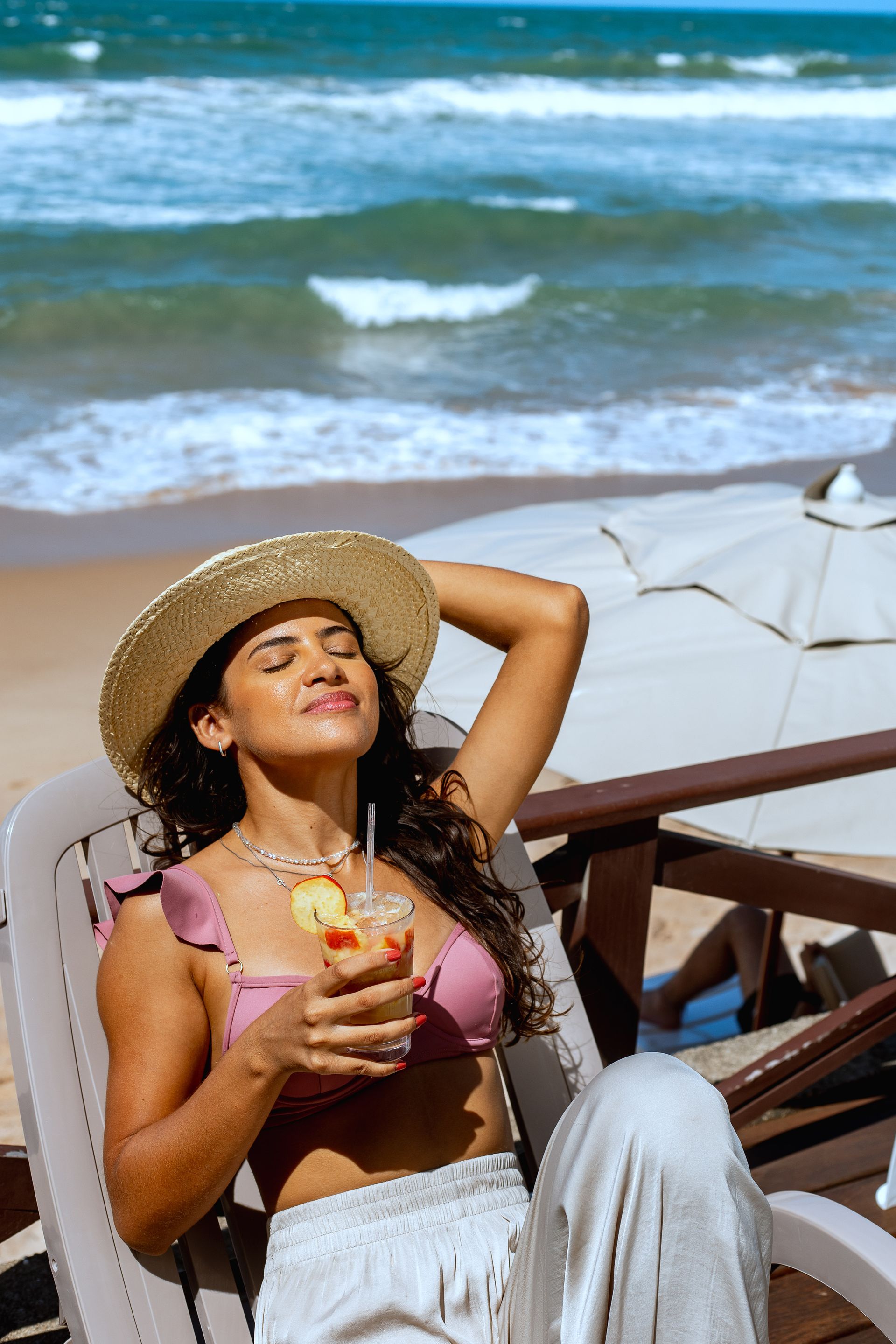 Mulher relaxando na praia, segurando uma bebida, usando chapéu e traje de banho. Oceano ao fundo.