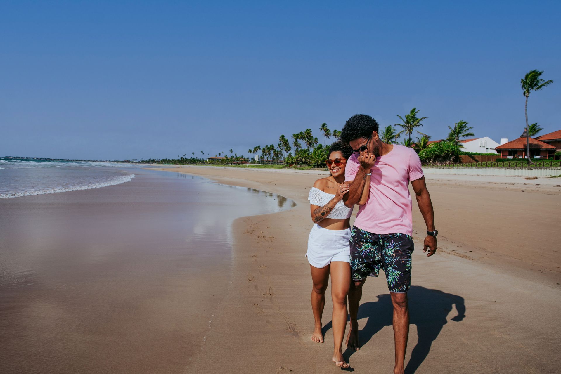 Casal caminha pela areia da praia. O homem veste uma camisa rosa, a mulher uma blusa branca e shorts. Céu azul no alto.